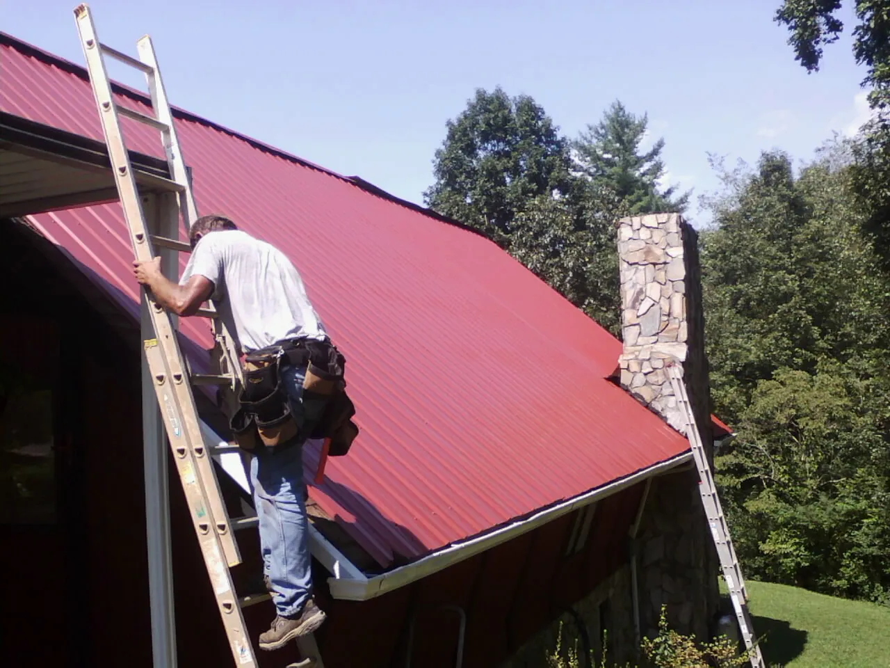 Roofer on a ladder beside a red metal roof and stone chimney — Soffit & Fascia Repair by Roofing Brothers in Grand Junction